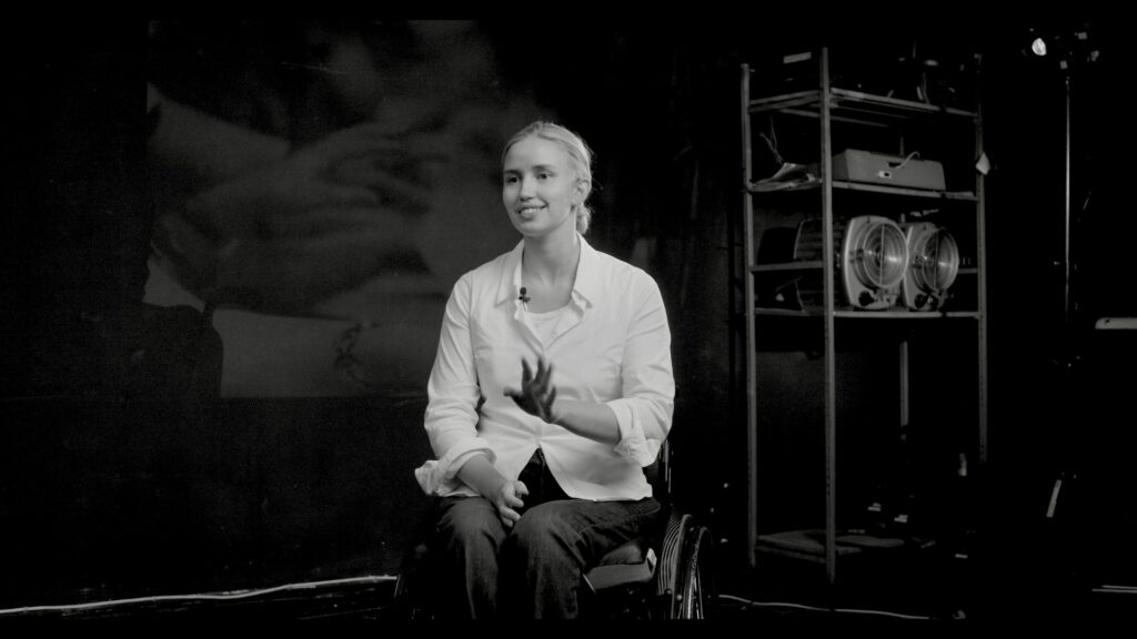 Portrait de Xénia Doroshenko, souriante, assise en fauteuil lors d’un entretien filmé dans un studio sombre. Elle parle avec les mains et regarde la caméra.