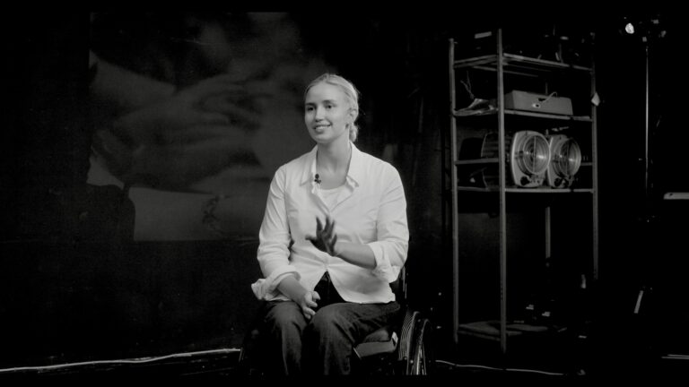 Portrait de Xénia Doroshenko, souriante, assise en fauteuil lors d’un entretien filmé dans un studio sombre. Elle parle avec les mains et regarde la caméra.