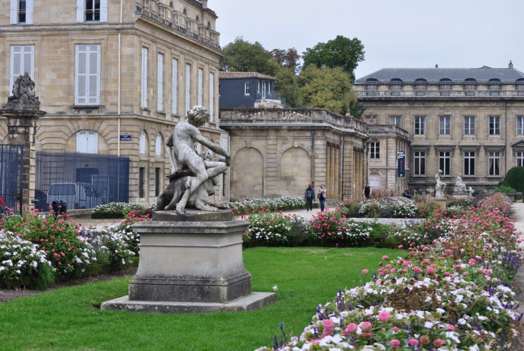 Statue de Jeunesse et chimère par Pierre Granet sur la terrasse Burguet, jardin public de Bordeaux, avec parterre fleuri et bâtiments du XVIIIe siècle.