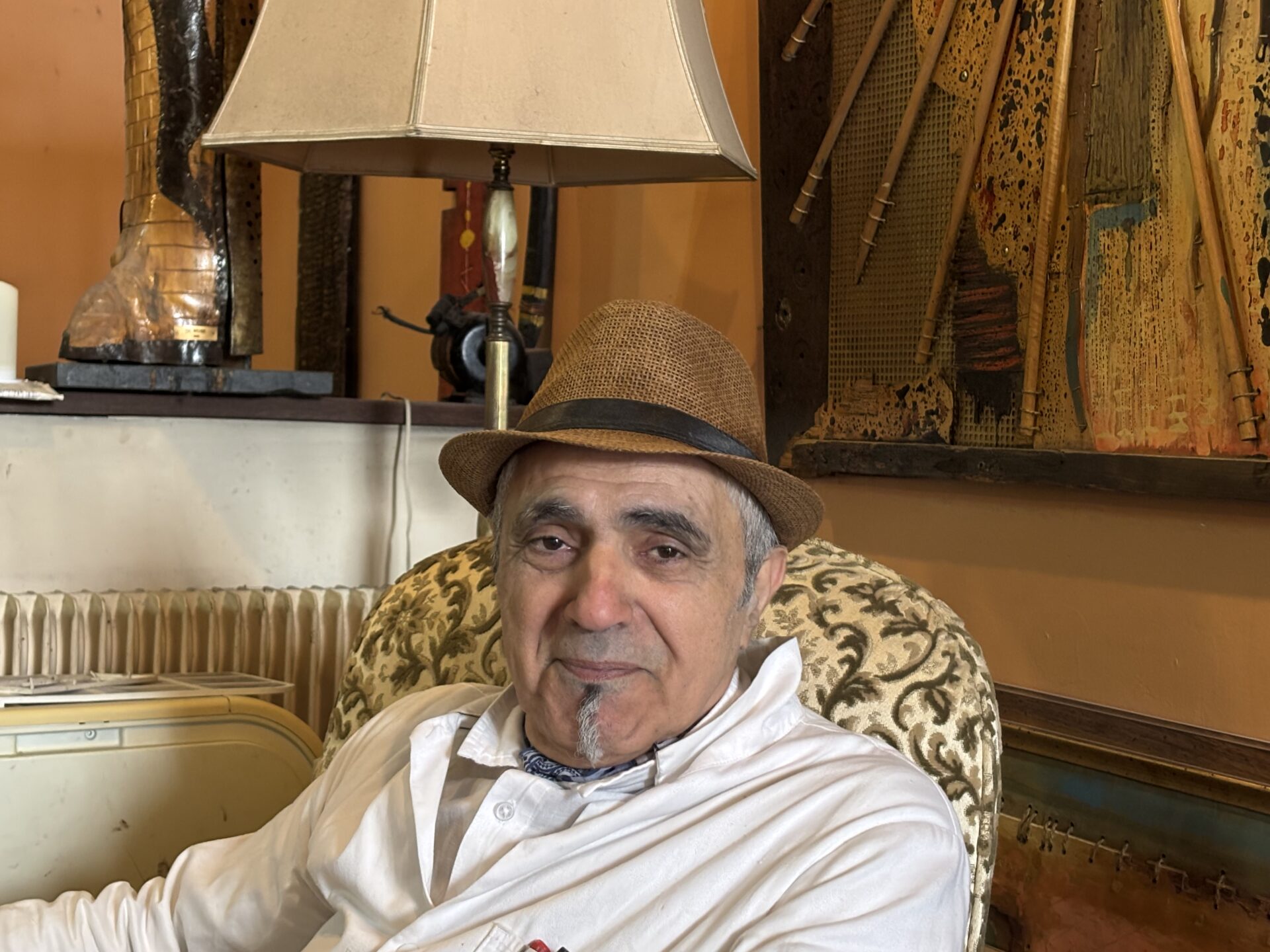 Maxime Veber assis dans un fauteuil, portant un chapeau, dans un intérieur chaleureux orné de tableaux et de sculptures